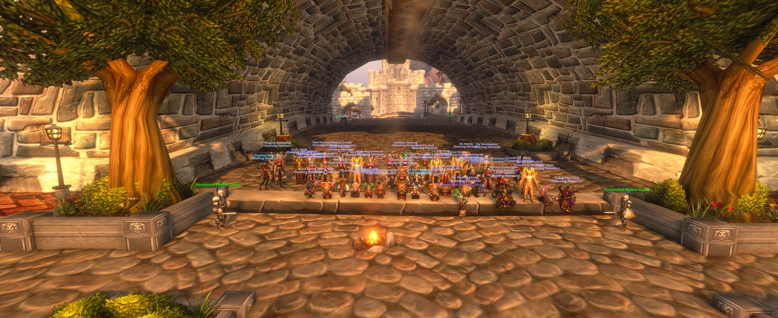 The guild lined up beneath a broad Stormwind stone arch, between trees and harbor guards, with a campfire in front.